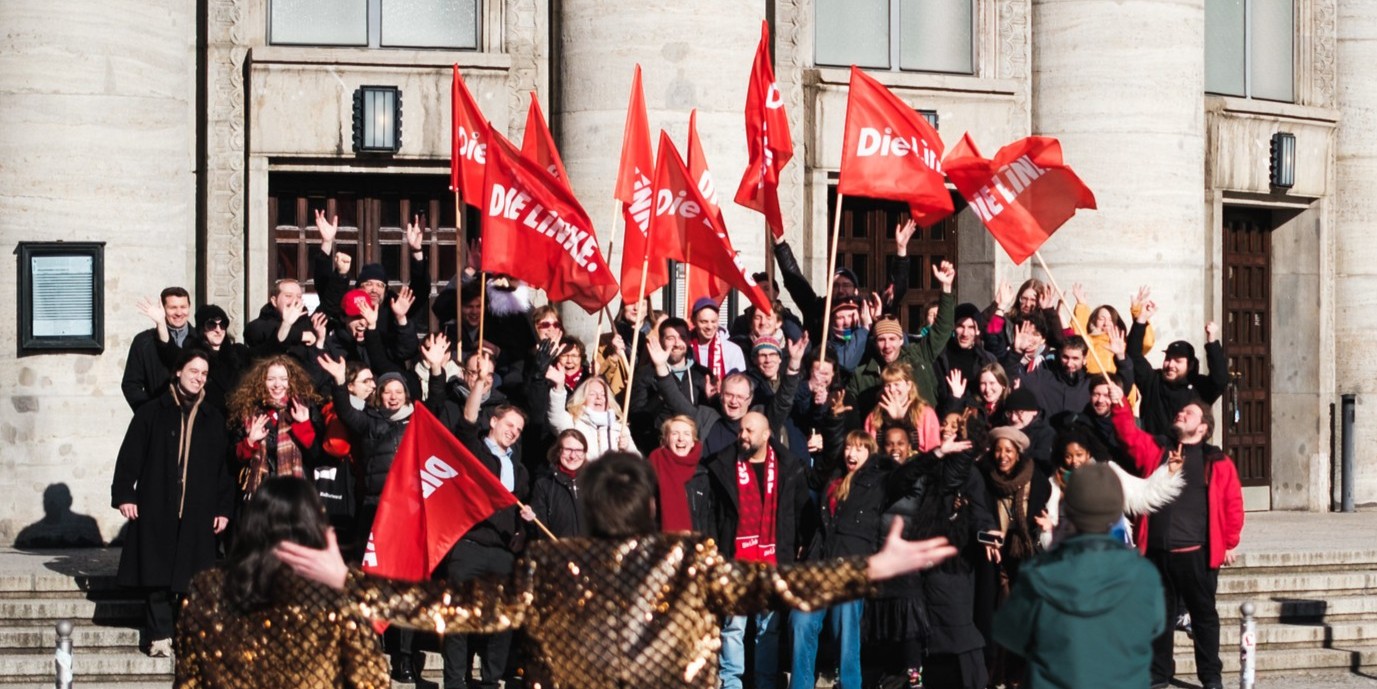 Partei-Header eine mittelgroße Gruppe von Menschen in winterlicher Kleidung steht auf den Stufen der Volksbühne. Sie sehen fröhlich aus und schenken rote Fahnen mit dem Logo der Partei Die Linke