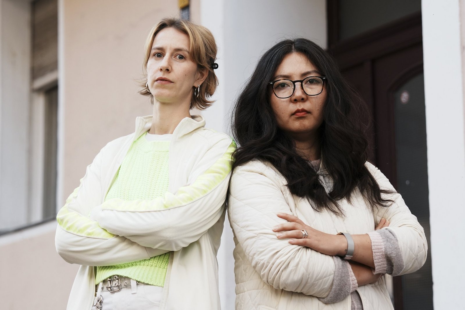 Zwei Frauen stehen mit verschränkten Armen, Rücken an Rücken, vor einem Hauseingang