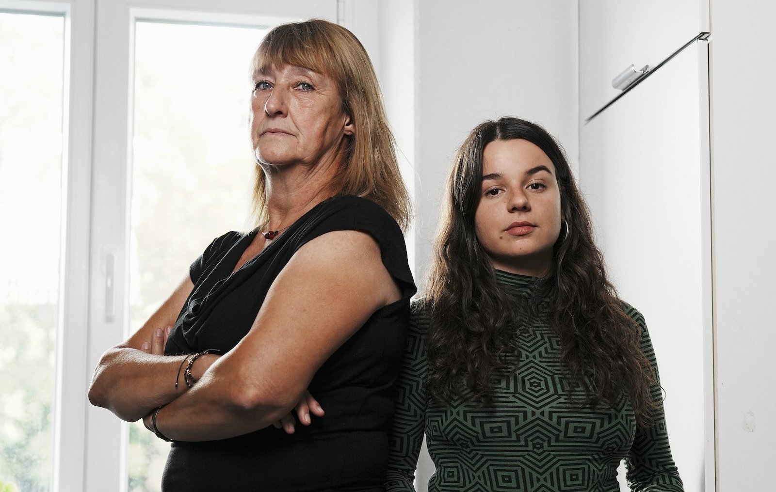 Zwei Frauen stehen nebeneinander in einem hellen Raum mit verschränkten Armen und ernstem Blick in die Kamera. Im Hintergrund ist ein Fenster mit Tageslicht zu sehen.