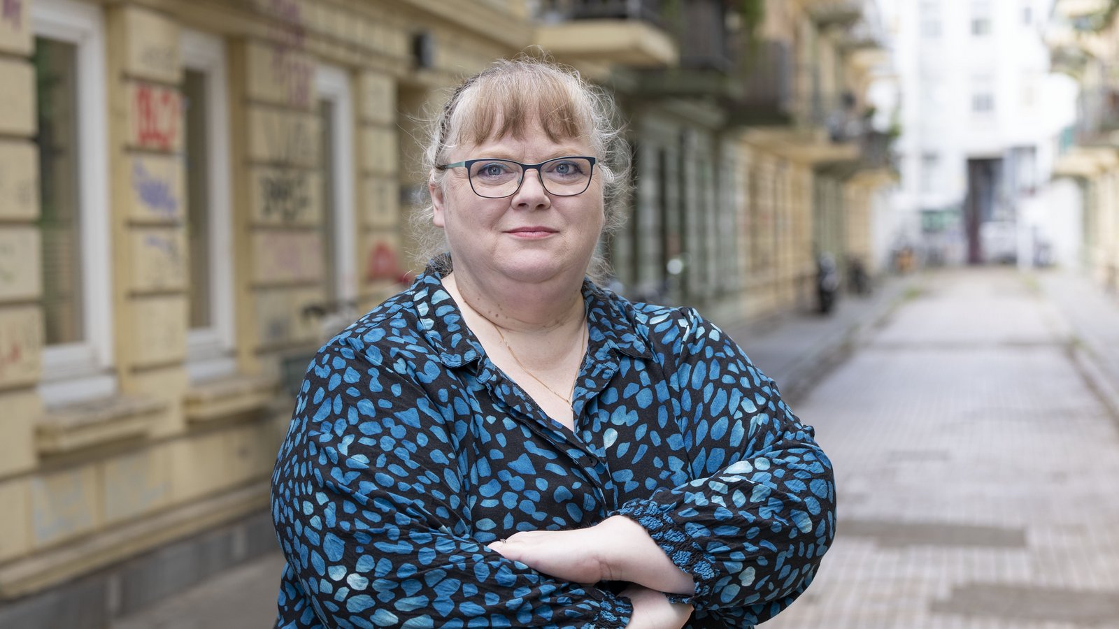 Olga Fritzsche trägt eine blaue Bluse und eine Brille. Sie steht mit verschränkten Armen vor einer Hausfassade.