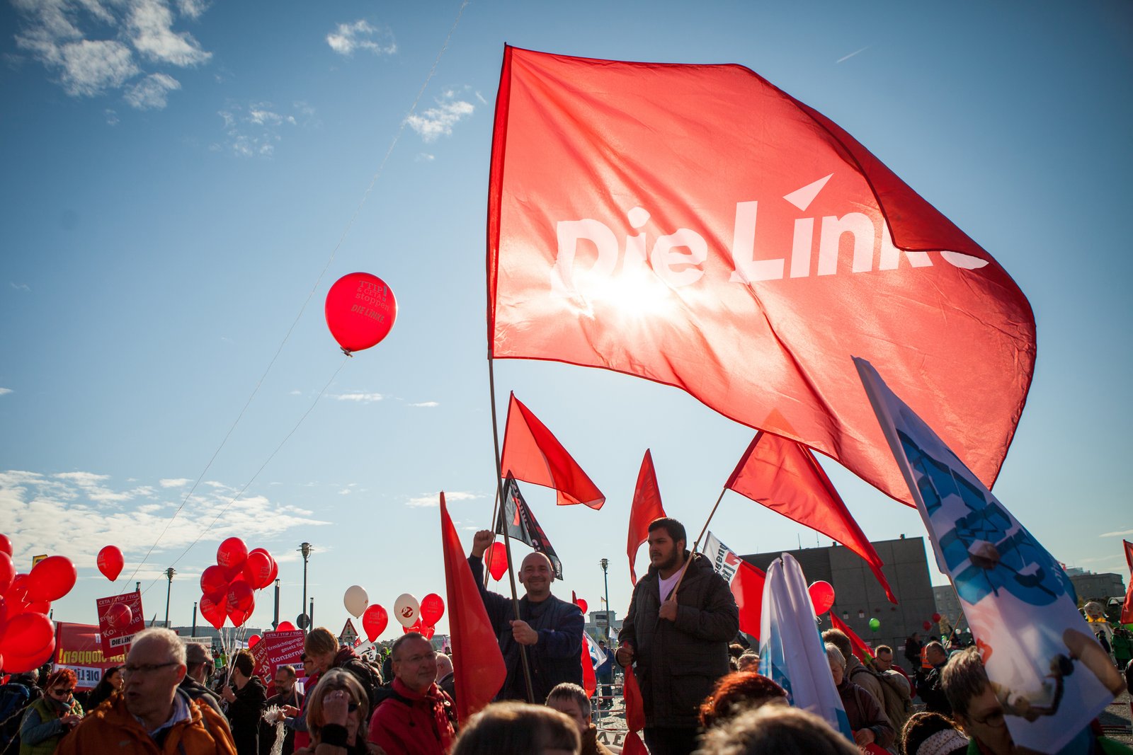 Im unteren Teil des Bildes ist eine Ansammlung an Menschen zu sehen. Sie schwenken rote Fahnen mit dem Aufdruck "Die Linke" und halten rote Luftballons. 